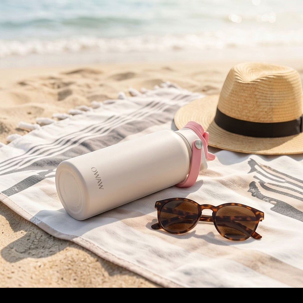 OWAW Nomade stainless steel water bottle in Blush Pink on a beach blanket with a sun hat